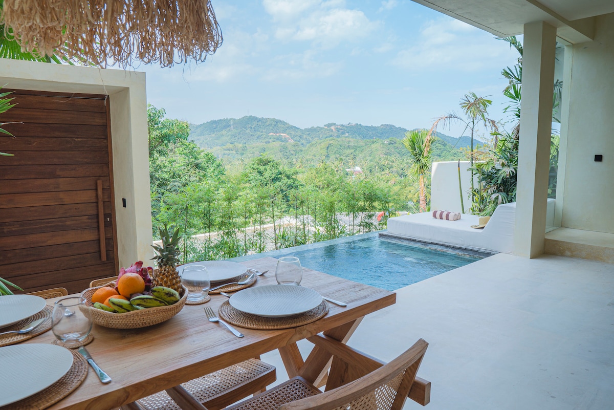 An outdoor dining area features a natural wood table set with plates and glasses, surrounded by tropical greenery. A private pool is visible nearby, reflecting the blue sky and mountains in the background. Lounge chairs are positioned near the pool for relaxation.