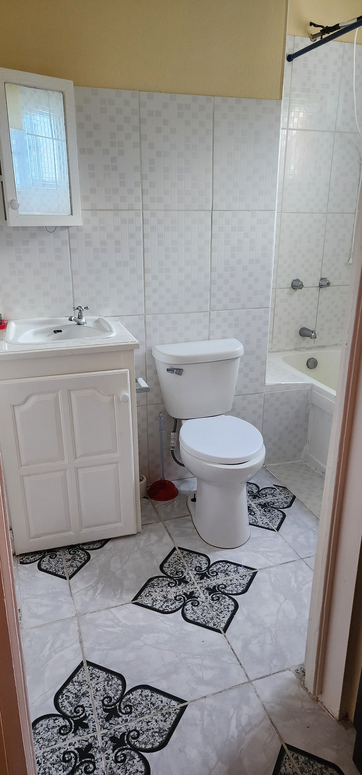 A compact bathroom is showcased, featuring a white vanity cabinet and a mirror. The space includes a toilet and a bathtub with a showerhead. White tiled walls create a clean aesthetic, while patterned flooring adds visual interest to the room.