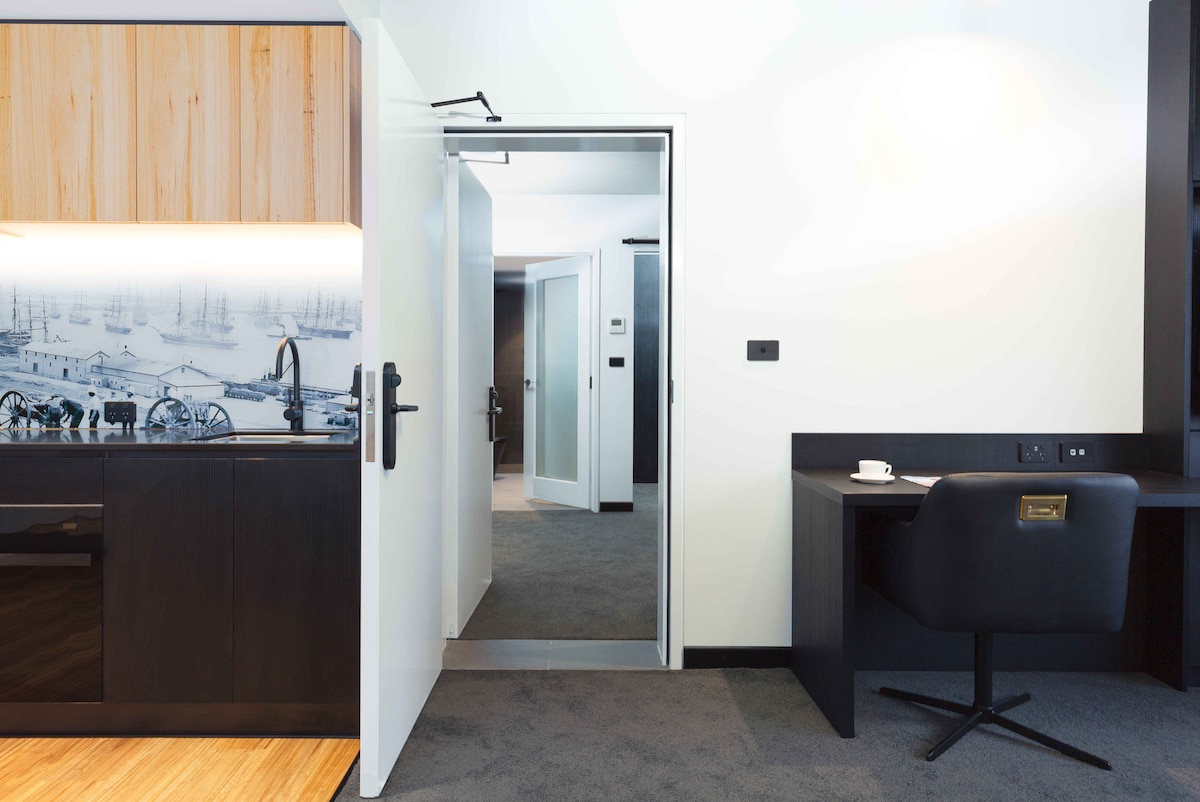 A well-appointed kitchenette is juxtaposed with a sleek black workspace. Soft lighting enhances the modern furnishings, while a door in the background provides access to the interconnecting apartment. Minimalist décor contributes to the open and airy feel of the space.