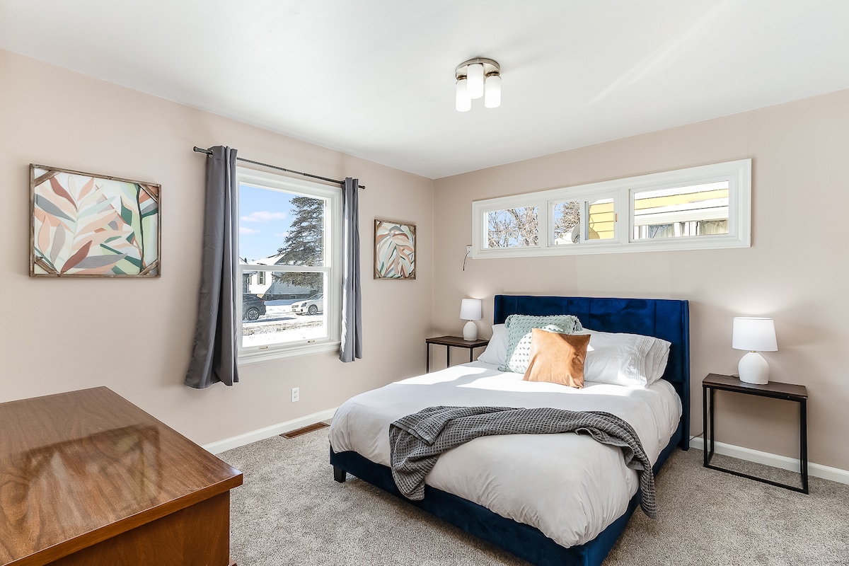 A bright bedroom features a spacious bed with soft linens, accented by a decorative pillow. Natural light enters through a window adorned with light curtains. Two nightstands with lamps flank the bed, and artwork hangs on the walls, adding a touch of color to the neutral decor.