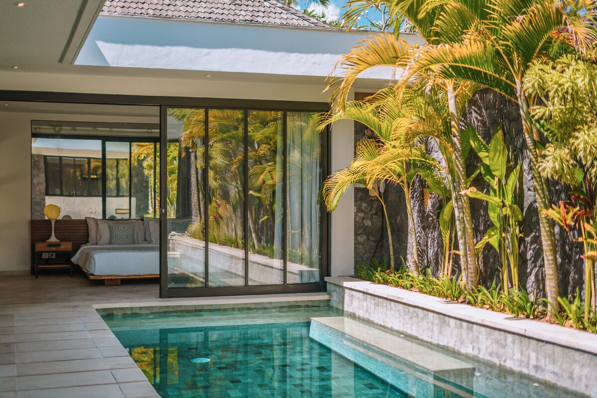 A minimalist living area opens to a tranquil private pool, surrounded by lush tropical plants. Large glass doors provide a seamless transition to the serene outdoor space, allowing ample natural light to brighten the interior. A cozy bed is visible in the background.