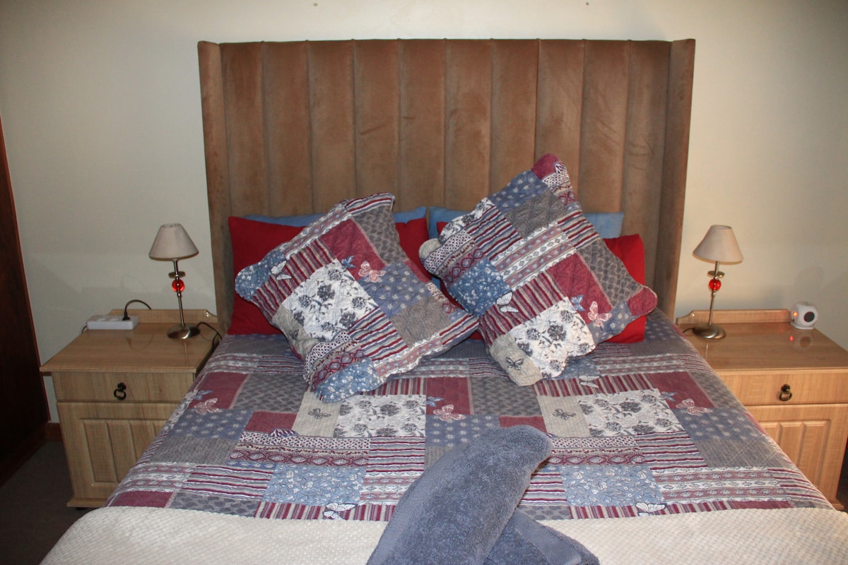A cozy bedroom is depicted with a spacious bed featuring a patchwork quilt and decorative pillows. Two bedside tables with lamps are positioned on either side of the bed, contributing to a comfortable and inviting ambiance.