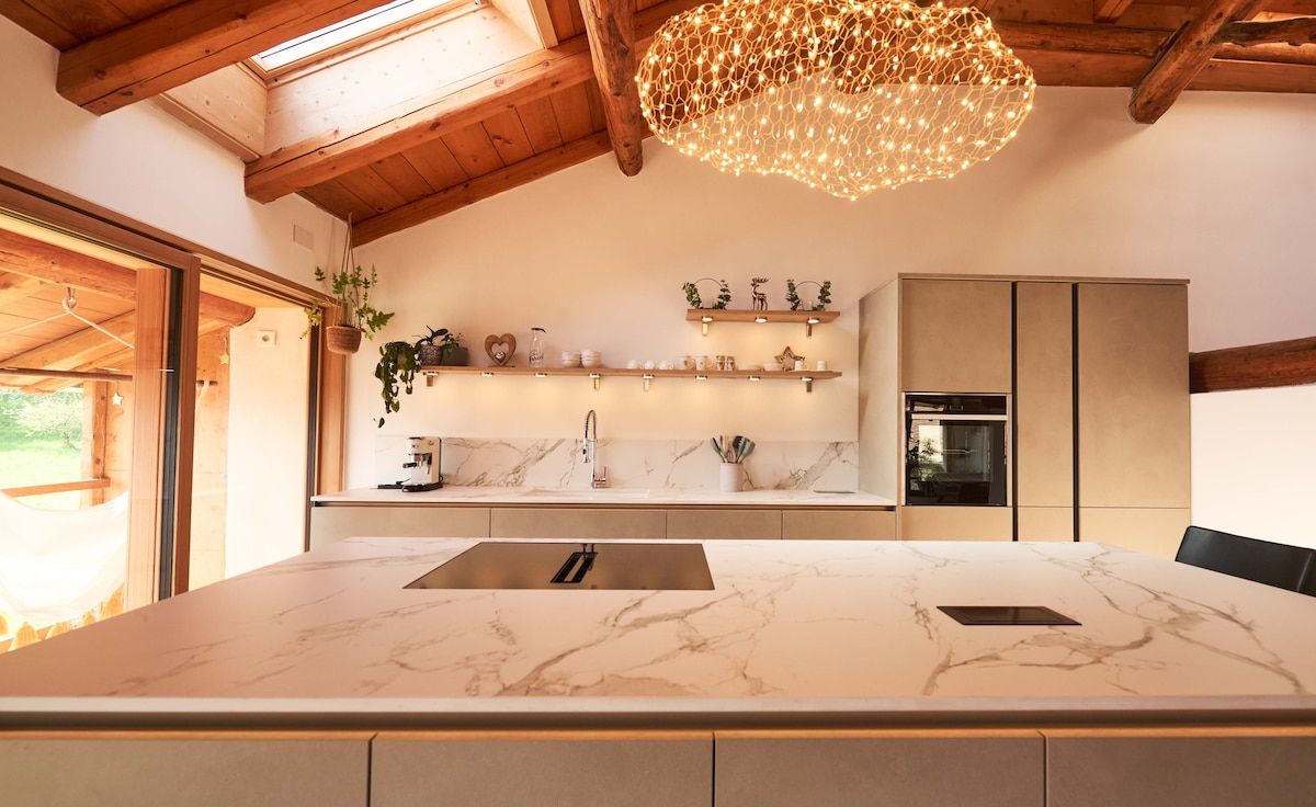 A modern kitchen features a large marble countertop with an integrated sink and stovetop. Open shelving displays decorative items. A striking light fixture made of small bulbs illuminates the space, complemented by wooden beams and natural light from the skylight.