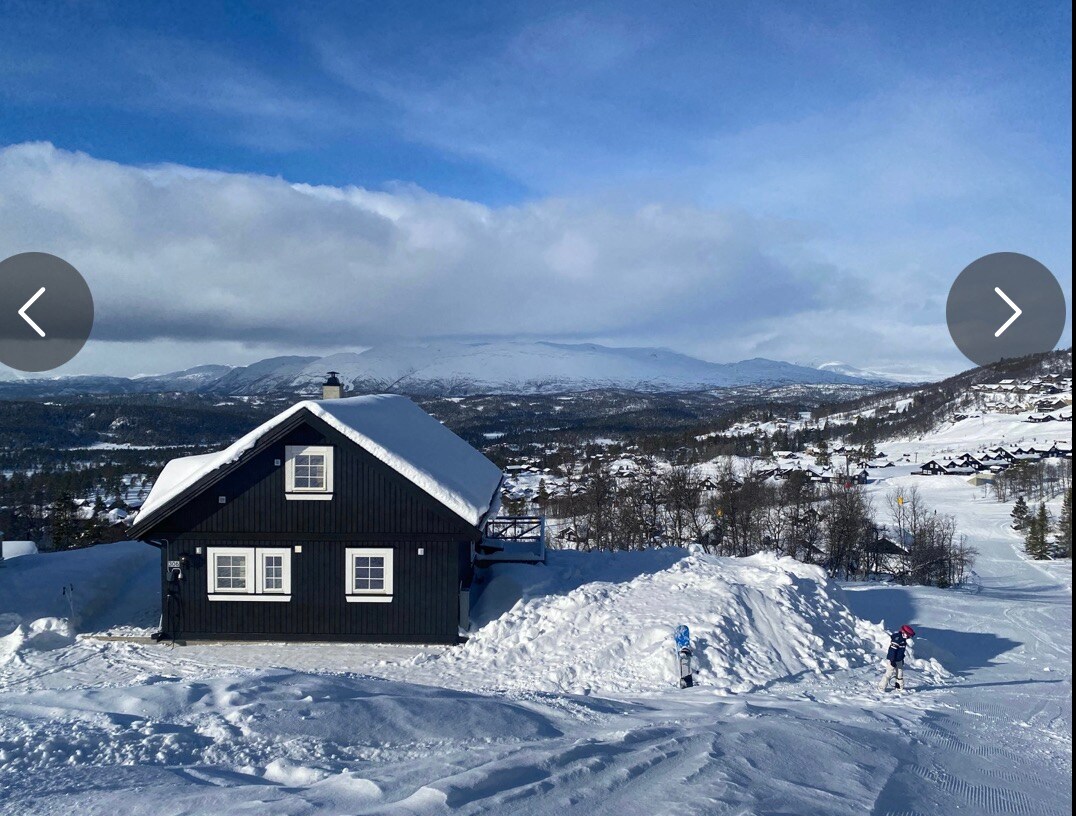 A black wooden cabin is set against a backdrop of snow-covered hills and blue skies. The surrounding landscape features a blanket of snow, with scattered trees and distant mountains visible. Tracks in the snow indicate paths leading away from the cabin.