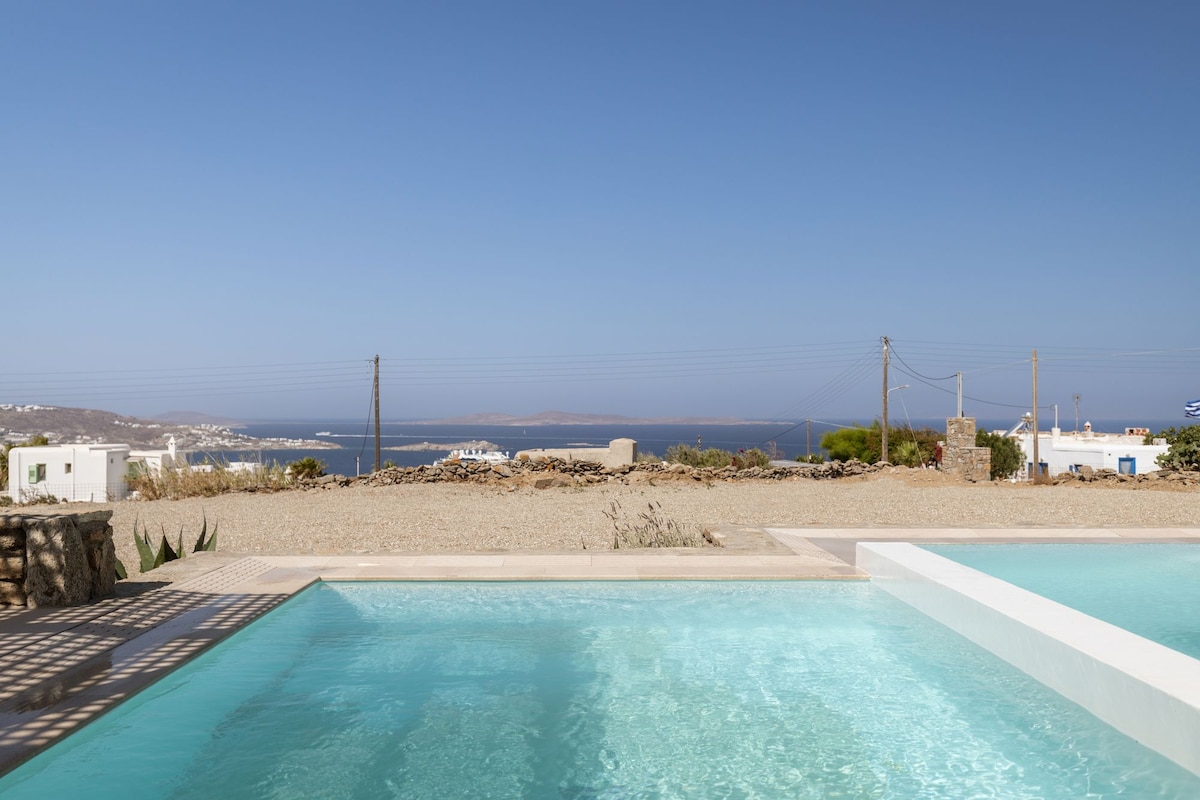 The image features a serene outdoor area with a clear swimming pool reflecting the sky. Beyond the pool, an expansive view of a coastal landscape and distant hills can be observed, while a stone wall frames the scene, adding a natural element.