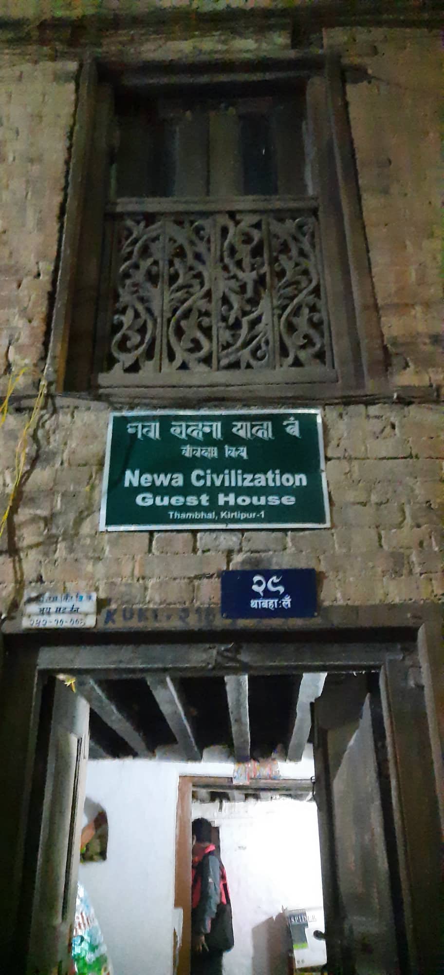 An entrance to Newa Civilization Guest House is showcased, highlighted by a wooden window with intricate carvings above a green sign displaying the name of the establishment. Textural details of the red-brick exterior and wooden beams are visible, reflecting traditional Newar architecture.