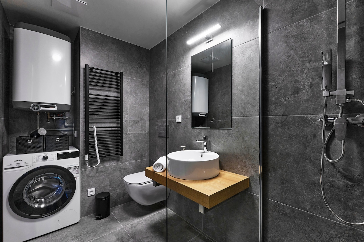 A modern bathroom features sleek grey tiles and minimalist fixtures. A circular sink is set on a wooden countertop, and a walk-in shower is enclosed with glass. A washing machine and a towel radiator are also present, maintaining a tidy and functional space.