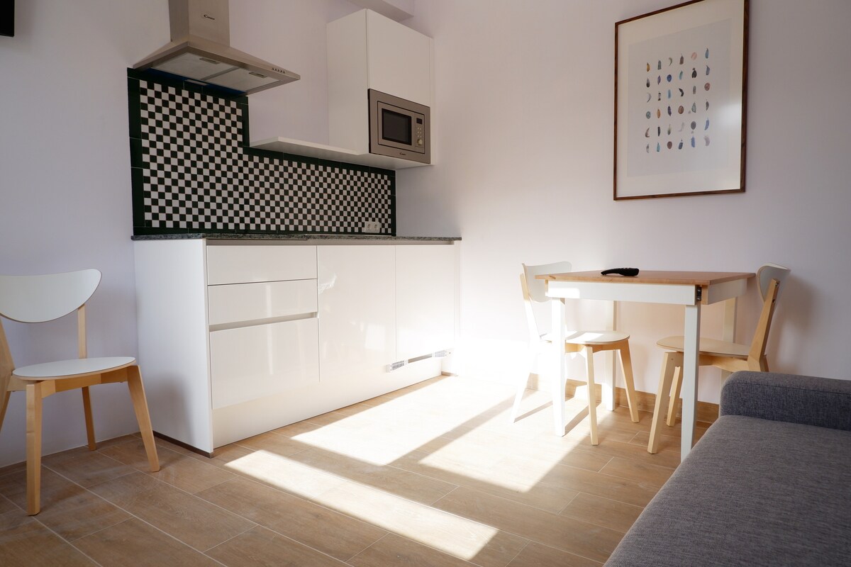 A modern kitchen area features sleek white cabinetry and a checkered backsplash. A table for two is paired with light wood chairs, while a gray sofa sits nearby. Natural sunlight streams in, illuminating the space and creating a bright atmosphere.