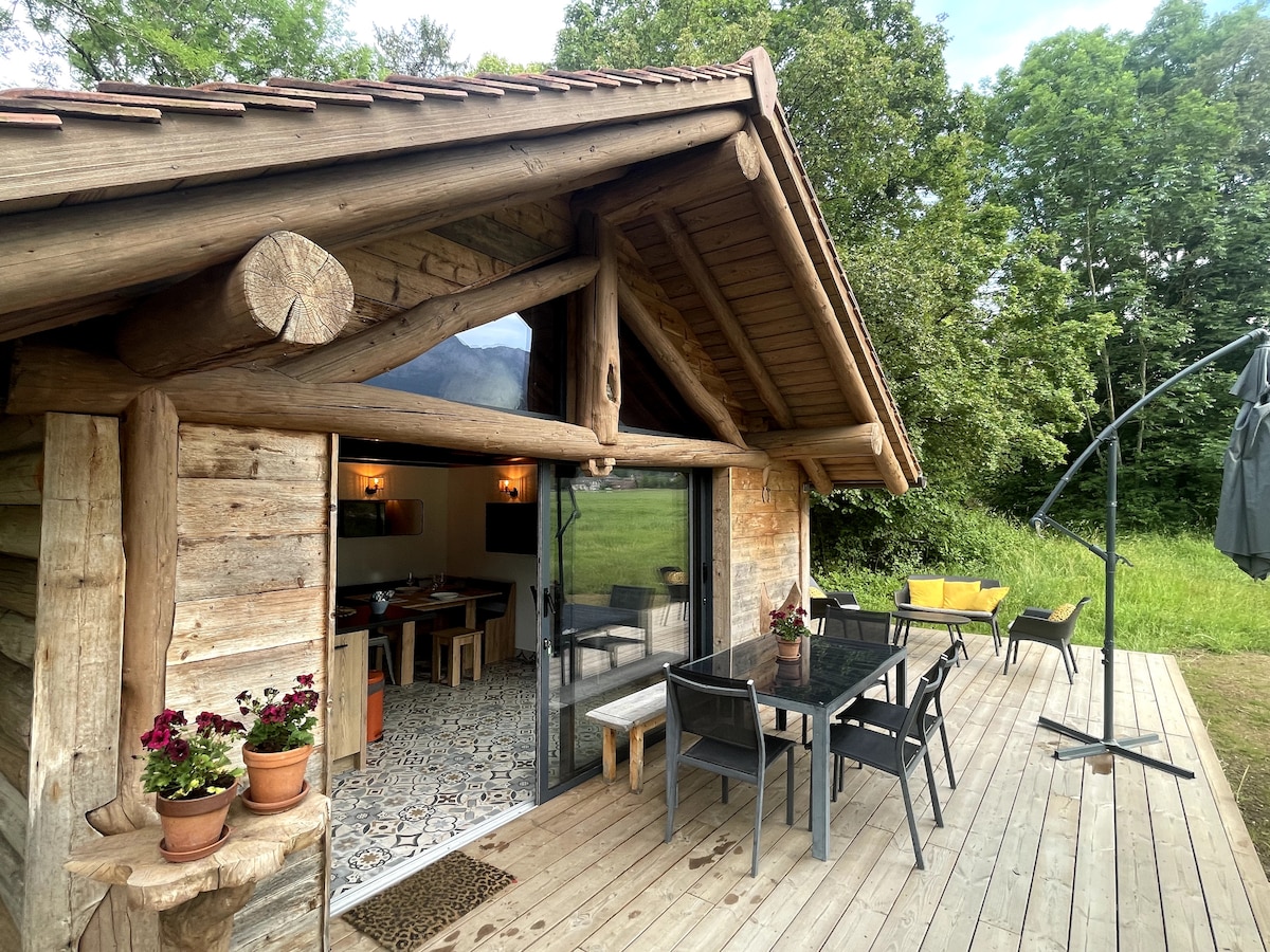 A wooden chalet exterior is displayed, with large glass doors opening onto a spacious deck. Outdoor furniture, including a dining table and chairs, are positioned under a covered area. Potted flowers add a touch of color, surrounded by green fields and trees.