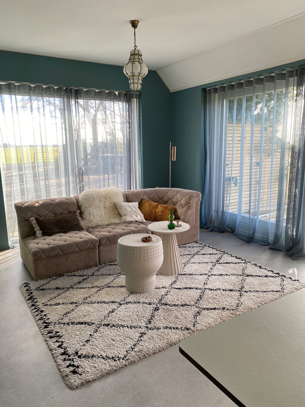 A cozy living area features a plush beige sofa accompanied by decorative cushions and a textured rug. Natural light streams through floor-to-ceiling sheer curtains, highlighting the open space. A small side table holds refreshments, creating an inviting environment for relaxation.