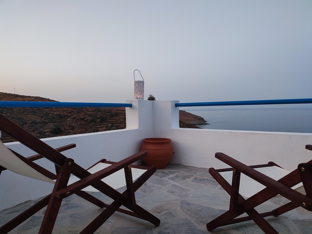 A spacious terrace features two wooden lounge chairs, positioned for relaxation. A clay pot sits in the center, complemented by a decorative lantern in the background. The calming blue railing offers views of the sea and coastal landscape, creating a serene outdoor space.