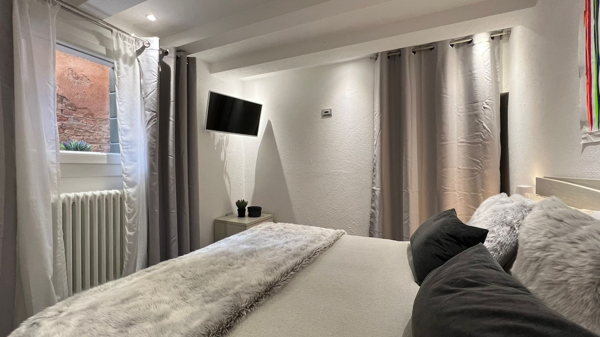 The bedroom features a spacious bed with gray pillows and a soft blanket. Natural light enters through the window, complemented by white curtains. A wall-mounted TV is positioned near the bed, and a small plant sits on a bedside table.