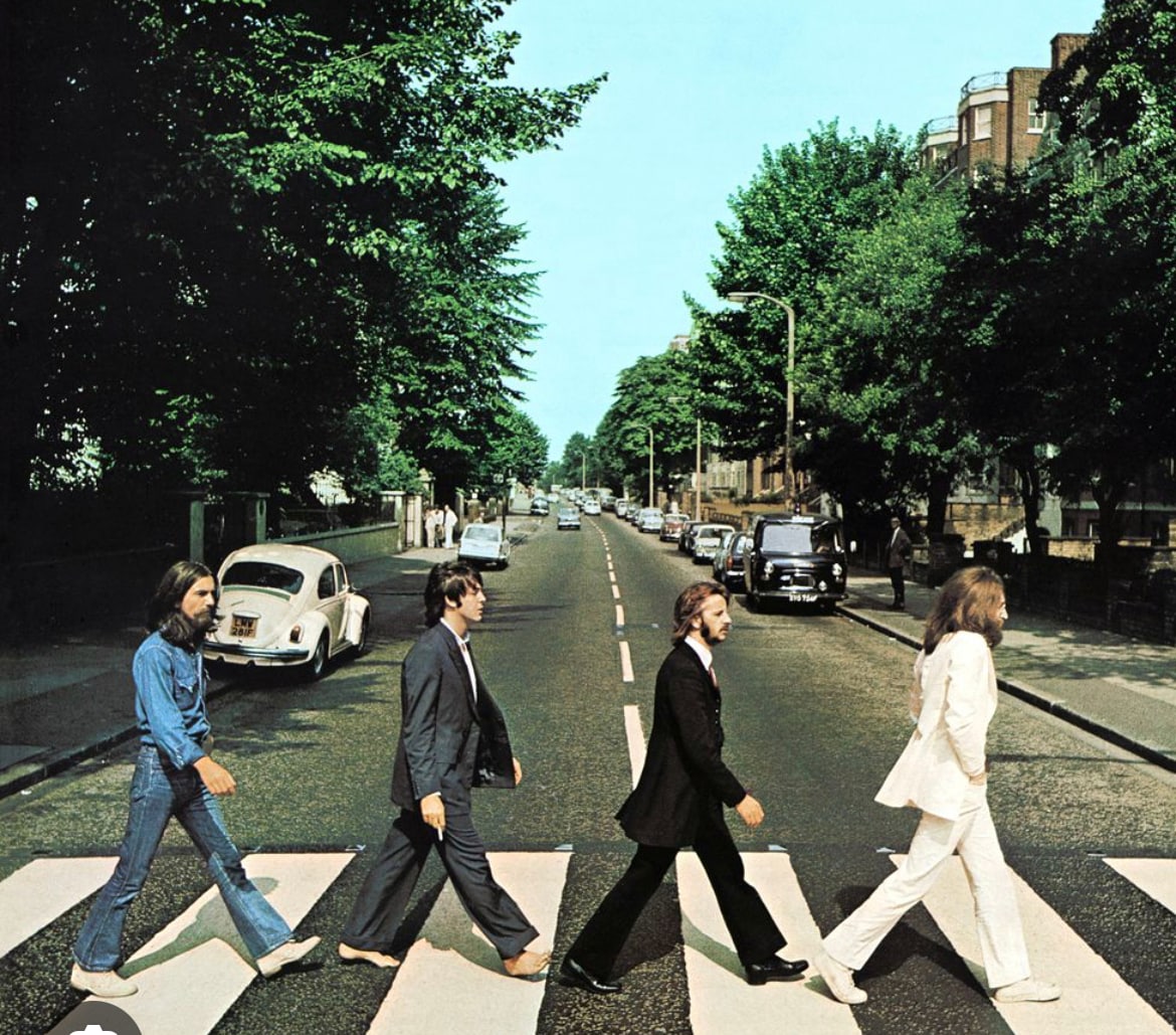 A wide street is seen with four individuals crossing at a marked pedestrian line. Vintage cars are parked along the sides, and tall trees line the street, adding greenery to the urban landscape. The clear blue sky enhances the scene's openness.
