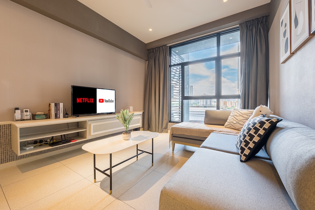 A modern living area is presented with a cozy sectional sofa and a minimalist coffee table. A flat-screen television mounted on the wall displays Netflix and YouTube logos. Large windows allow natural light to brighten the space, framed by elegant gray curtains.