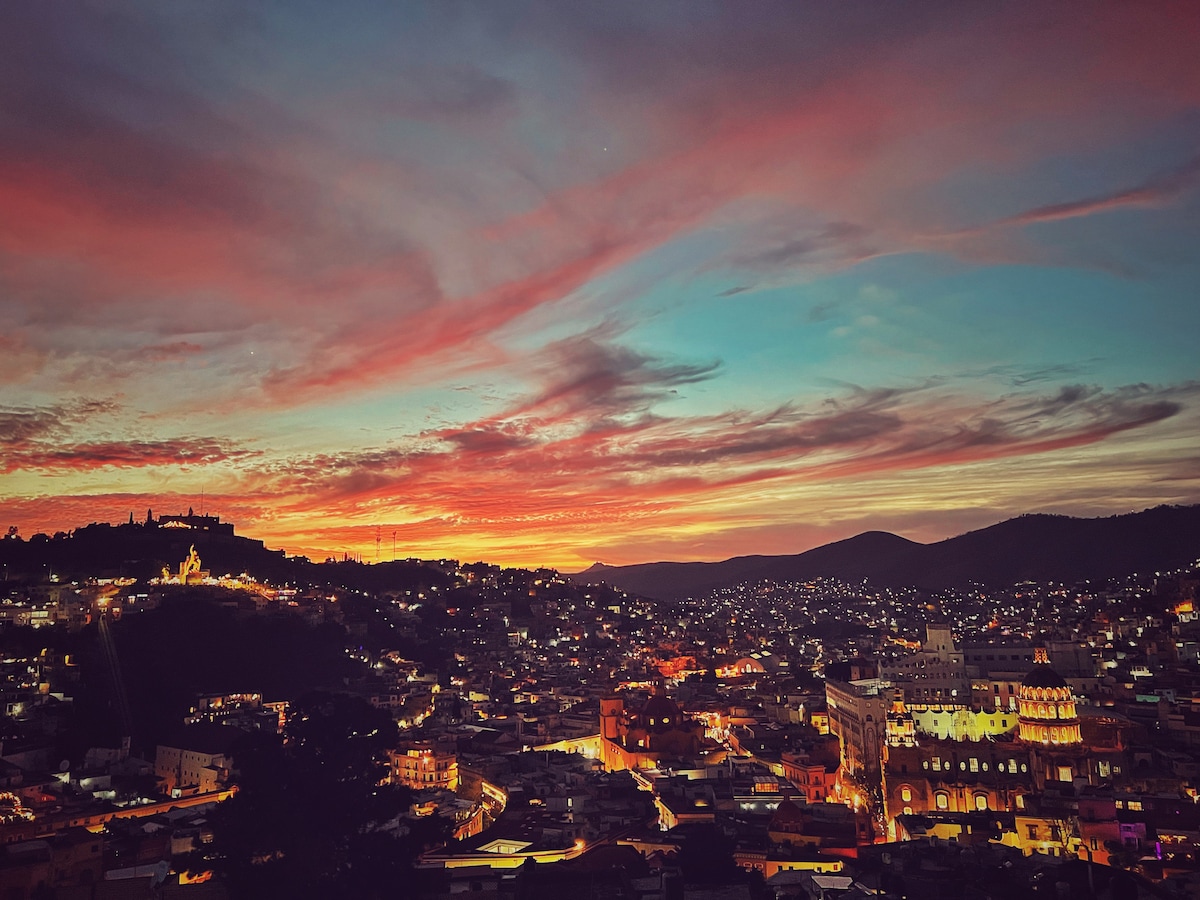 A vibrant sunset casts rich colors across the sky, transitioning from deep orange to soft blue. The view reveals a sprawling city below, with twinkling lights dotting the hillside, highlighting the unique architecture of Guanajuato against the dramatic backdrop of mountains.