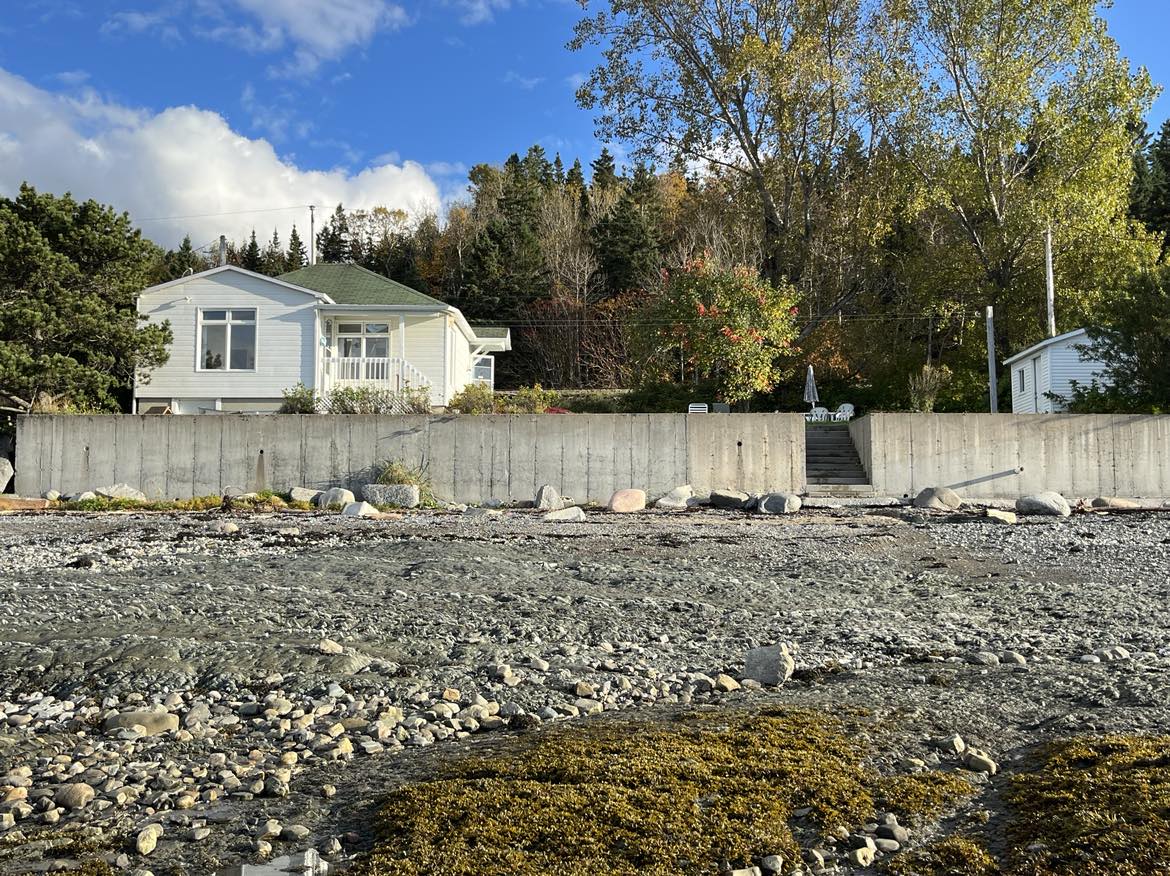 A charming beachfront property is seen across a rocky shore, framed by greenery and autumn trees. A concrete wall forms a boundary, with steps leading up to the residence. The clear sky highlights the serene atmosphere of the surrounding landscape.