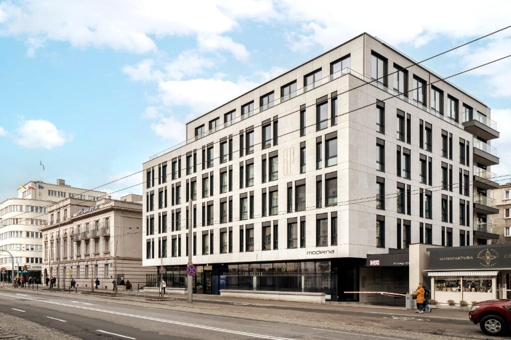 The modern building features a clean, minimalist design with large windows and balconies on the upper floors. The surrounding area includes various shops and buildings, contributing to the vibrant city atmosphere. This three-story structure showcases a blend of contemporary architecture with functional spaces.