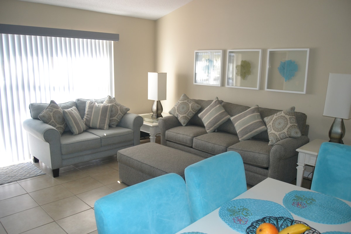 The living area features a comfortable gray sofa and chair adorned with patterned cushions. A coffee table sits between them, and natural light streams through vertical blinds. An adjacent dining area is highlighted by a bright table set with turquoise accents and fresh fruit.