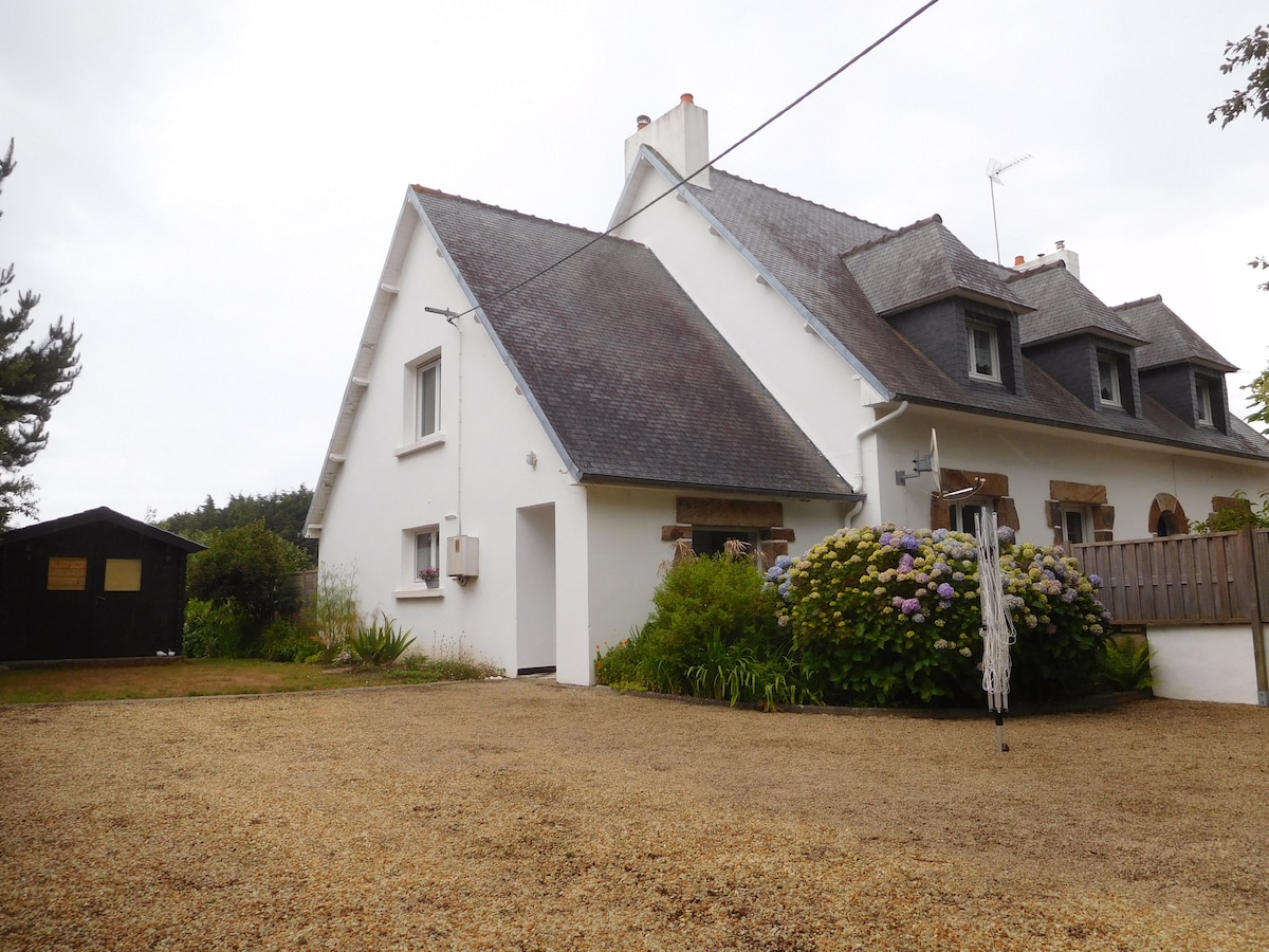 The exterior of a two-story neo-Breton house is visible, featuring a sloped roof and a light-colored facade. Surrounding greenery and colorful flowers add to the setting. A gravel driveway leads to the entrance, and a small garden shed can be seen in the background.