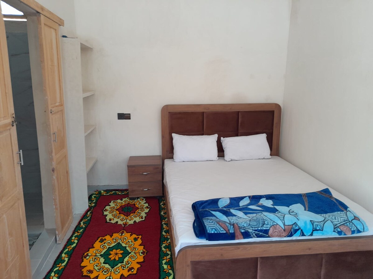 A simple bedroom features a spacious bed with two white pillows and a patterned blanket. A vibrant rug adds color to the floor. Nearby, a wooden bedside table and a set of shelves provide practical storage. A door leads to an adjacent bathroom.