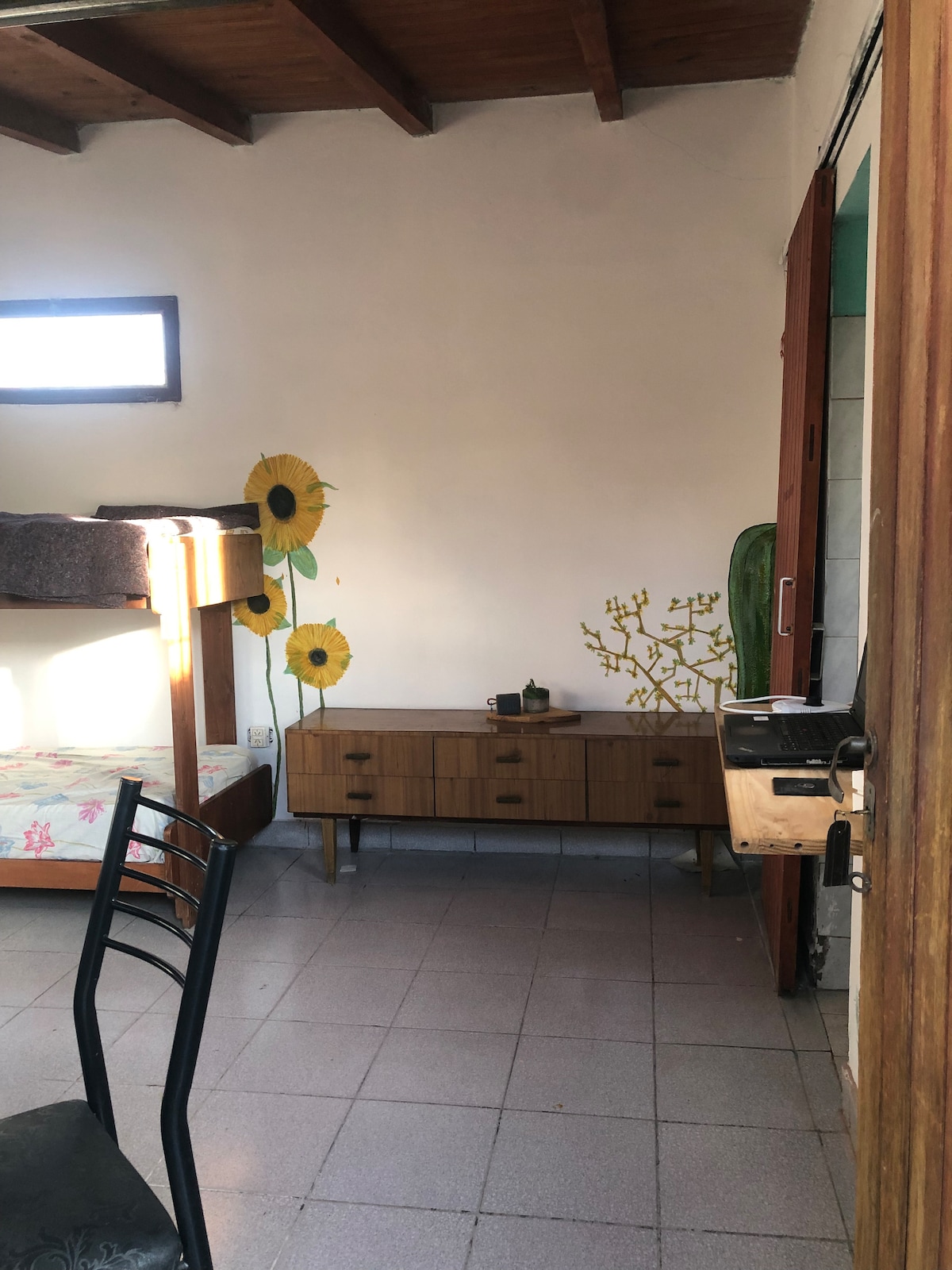 A cozy room features a set of bunk beds against a wall adorned with cheerful sunflowers. A wooden sideboard with a few decorative items is positioned beside the bed. Natural light enters through a window, illuminating the tiled floor and enhancing the room's airy feel.
