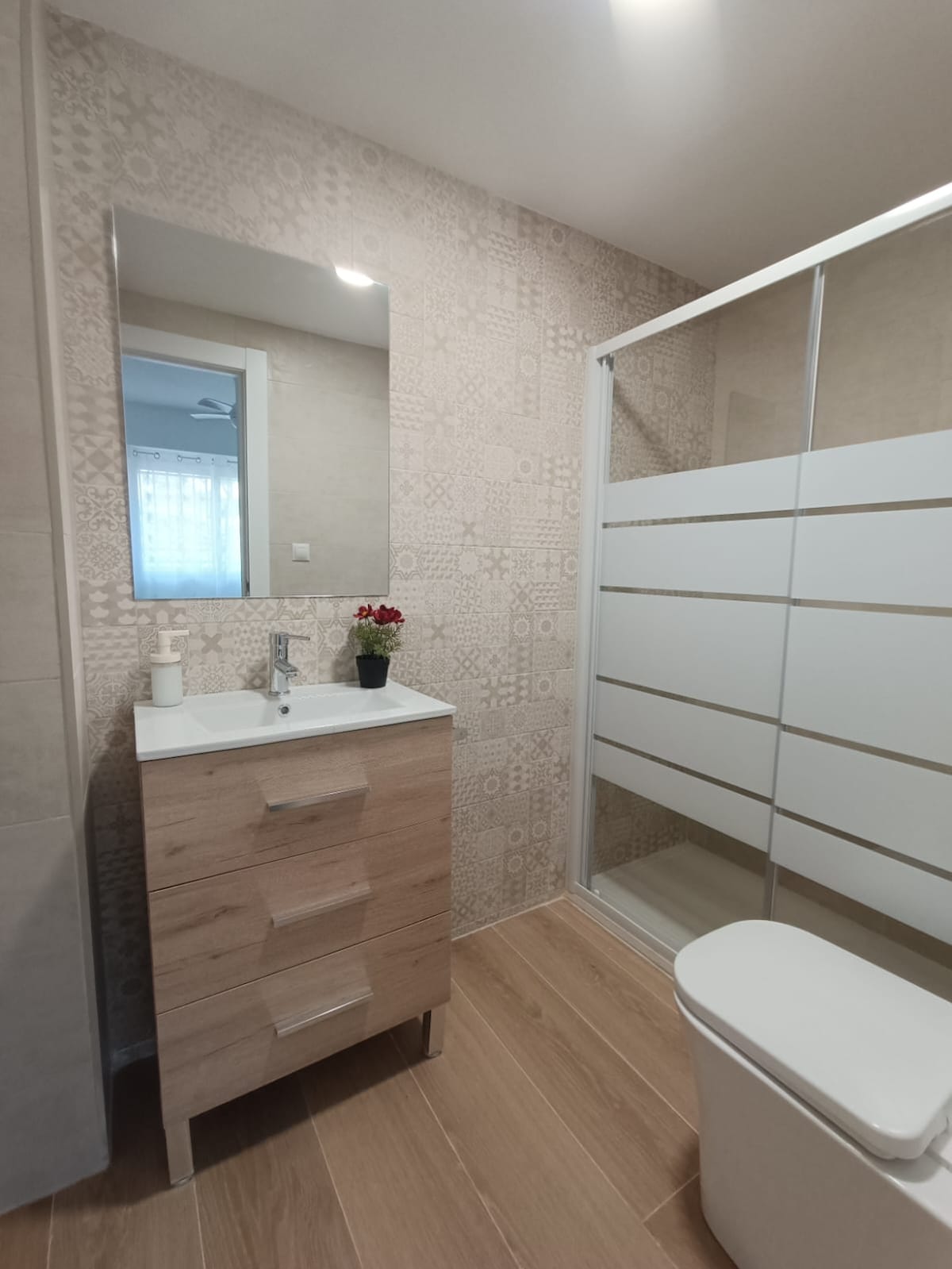 A modern bathroom is presented, featuring neutral-toned walls and warm wood accents. A shower with glass doors is visible alongside a sleek vanity unit with storage drawers. A small basin is situated above the countertop, complemented by a decorative plant. Natural light brightens the space.