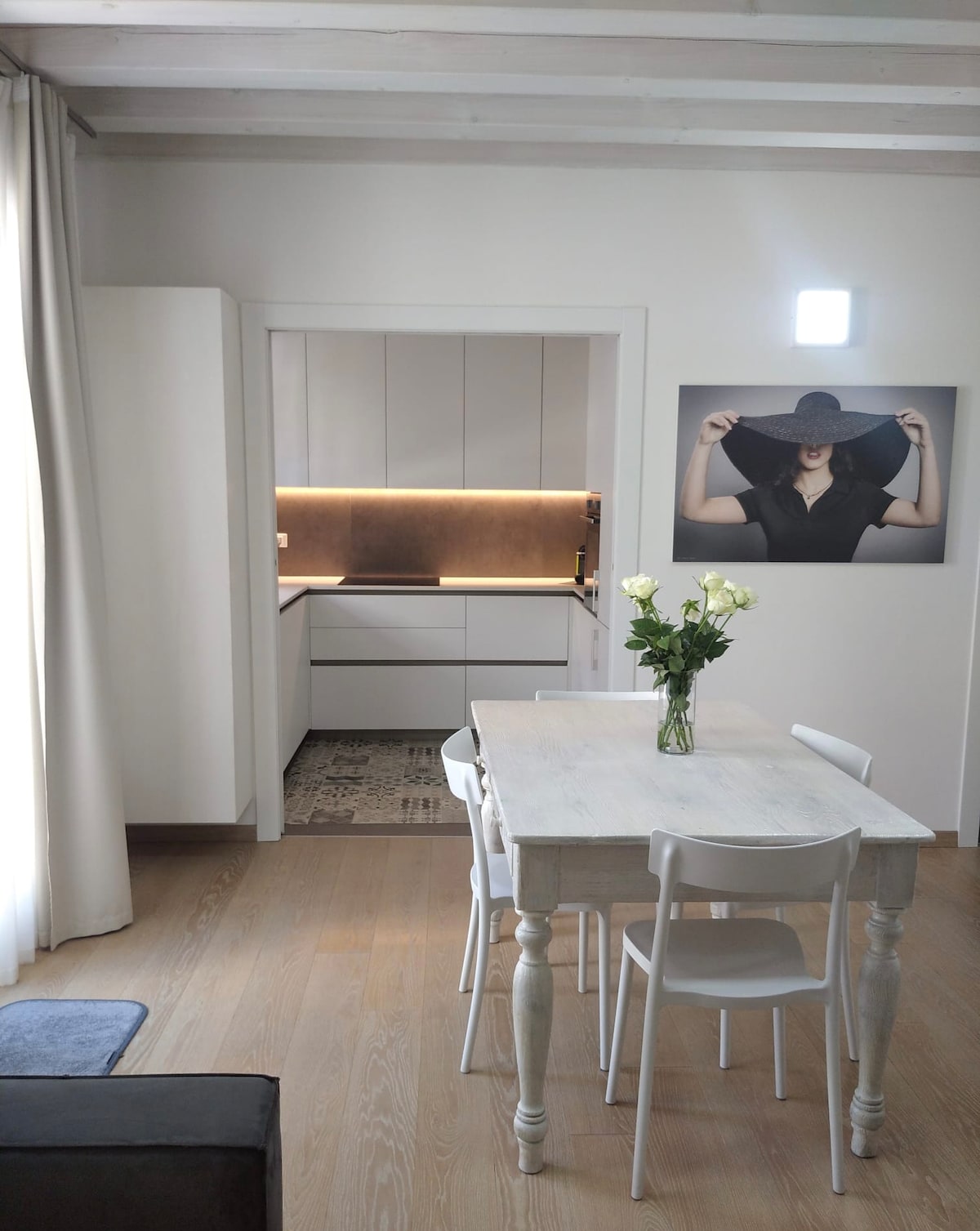 A modern kitchen area is visible through an open doorway, featuring sleek cabinetry and a subtle warm lighting. A white dining table with four chairs is positioned in the foreground, adorned with a vase of fresh roses. A large artwork is displayed on the wall adjacent to the kitchen.