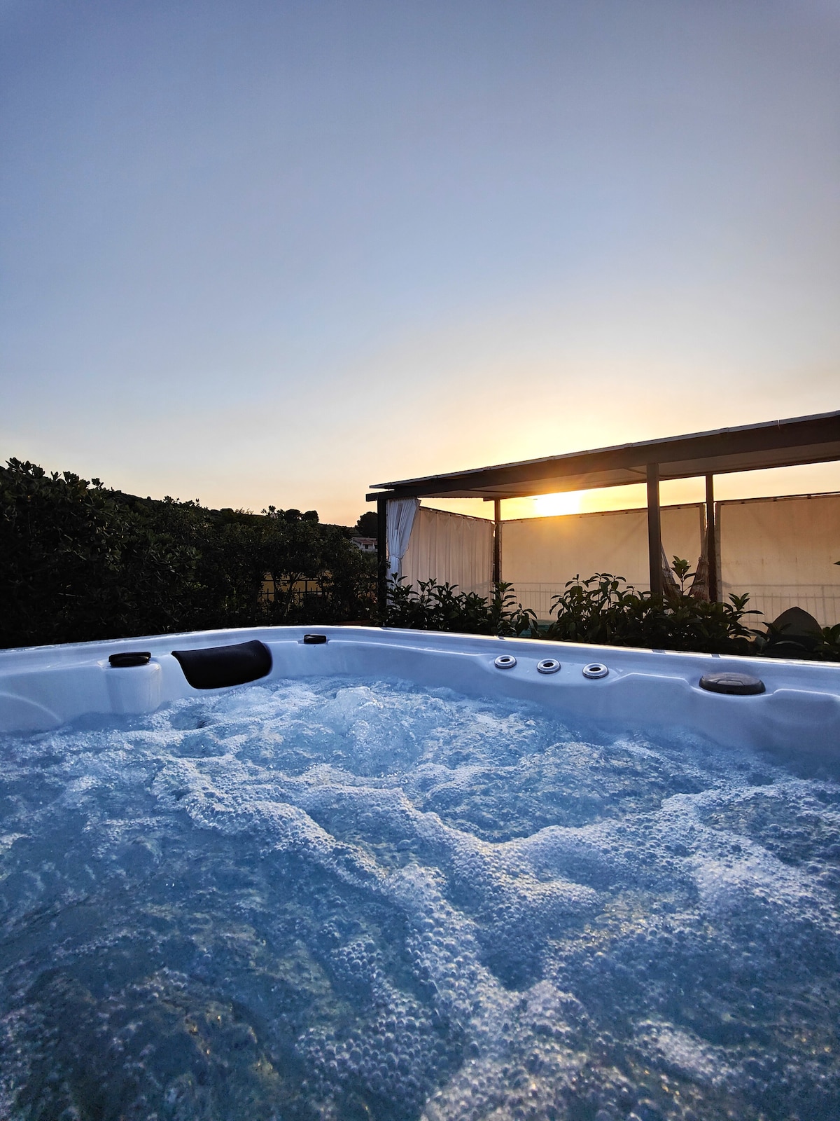 The image captures a relaxing hot tub with bubbling water, surrounded by lush greenery. The sun sets in the background, casting a warm glow over the scene, while a covered patio is visible nearby, enhancing the serene atmosphere.