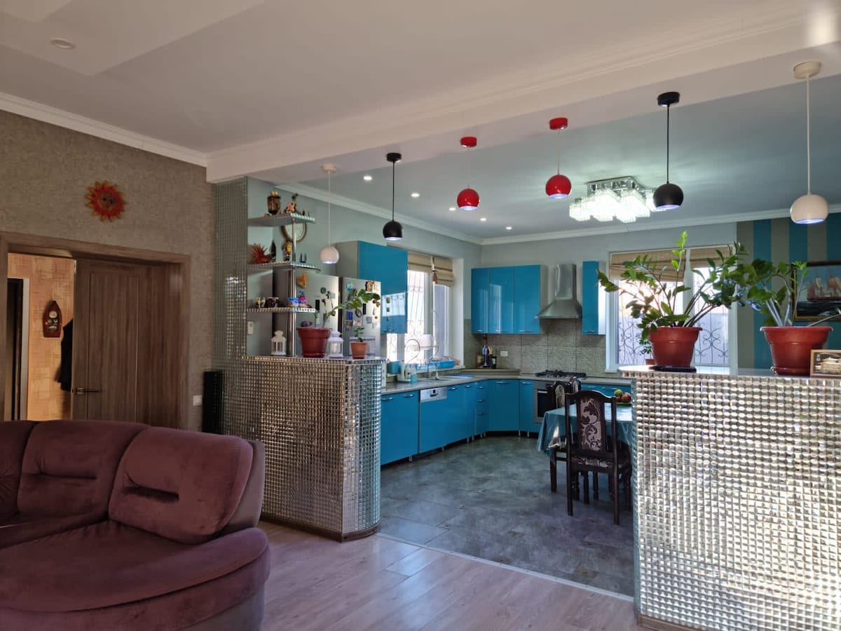 The kitchen area features vibrant blue cabinetry and stainless steel appliances. A mosaic, mirrored wall complements the modern design. Potted plants add a touch of greenery, while pendant lights in red and black provide gentle illumination over the dining space, creating a cozy atmosphere.
