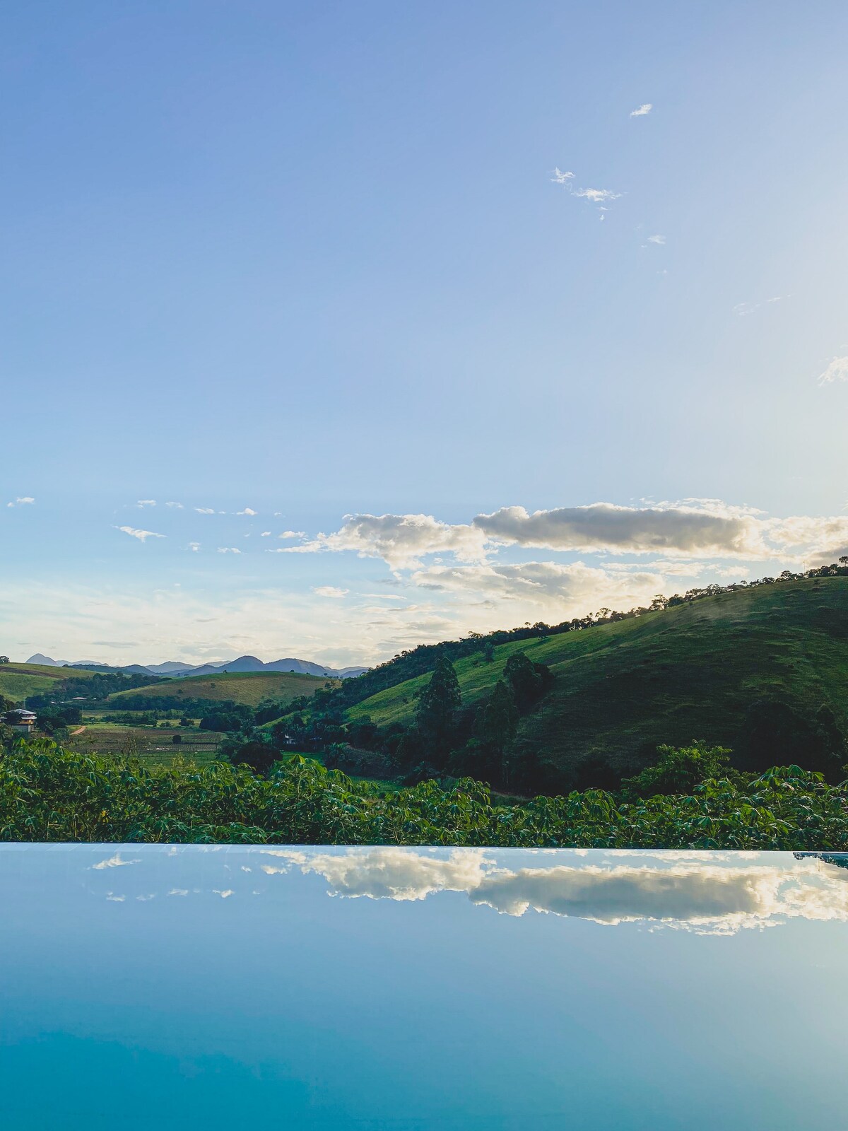 A panoramic view of rolling green hills is captured under a clear blue sky. Soft clouds float above, reflecting in the calm water of a nearby pool, creating a serene atmosphere that connects with the surrounding natural landscape.