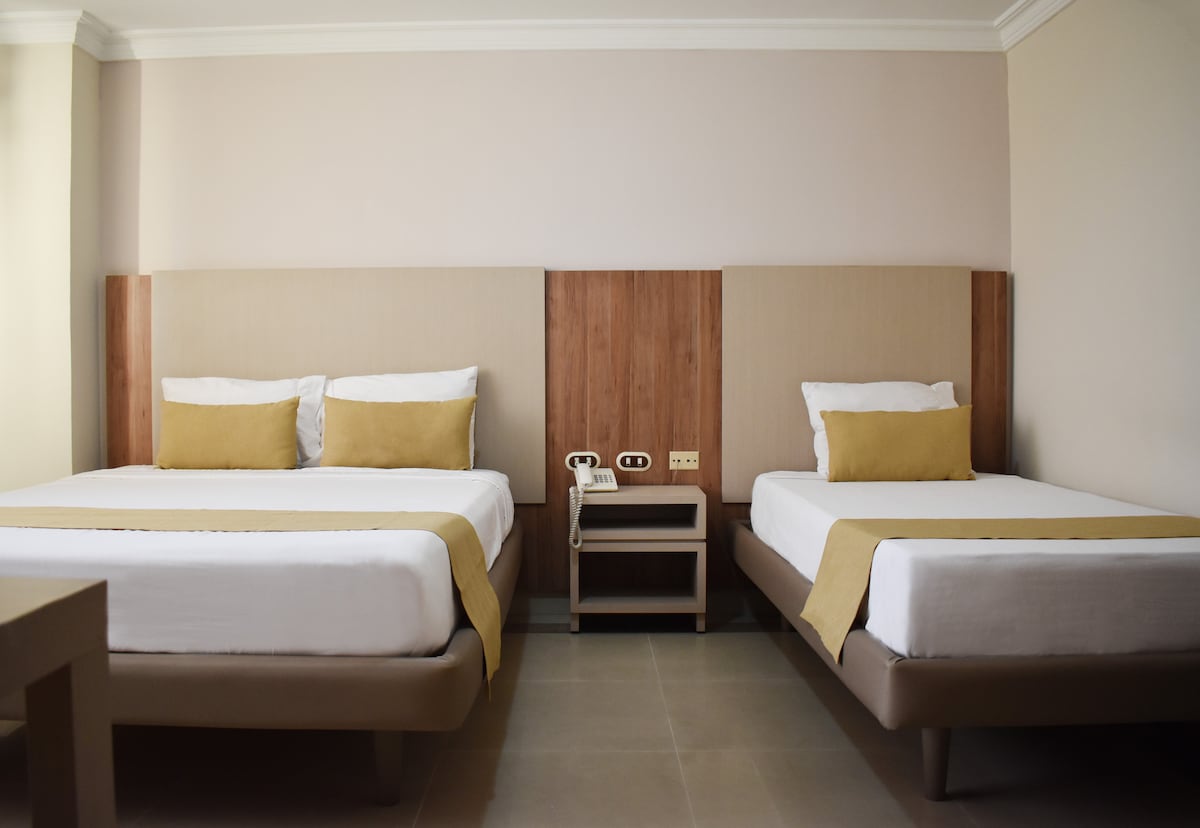 A comfortable guest room features two spacious beds, each with neutral-colored bedding and decorative throw pillows. A nightstand is positioned between the beds, displaying a telephone and power outlets. Soft beige walls complement the inviting design, creating a restful atmosphere.