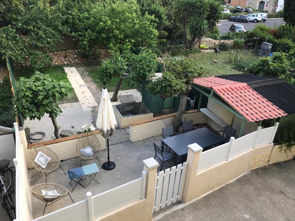 The garden area is viewable from above, featuring a shaded terrace with outdoor seating and a table. Lush greenery surrounds the space, with a pathway leading through the garden. A shaded area equipped with a grill is also visible, along with a small storage shed.