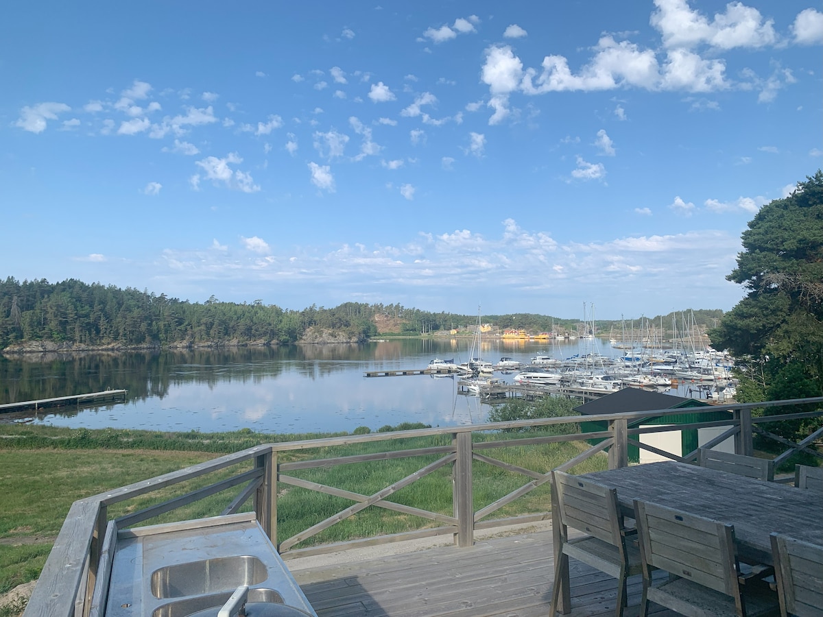 A serene view of a calm marina is presented, with multiple boats docked in the water. The scene features lush greenery in the background and a clear blue sky dotted with clouds. A wooden deck with a dining area is visible in the foreground.