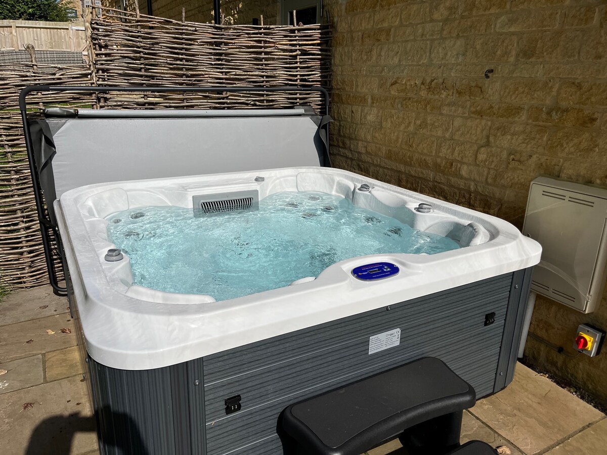 The image features a modern hot tub with bubbling water, positioned on a patio area. Natural stone walls serve as a backdrop, and the outdoor space is surrounded by a woven privacy screen, creating a serene atmosphere for relaxation.