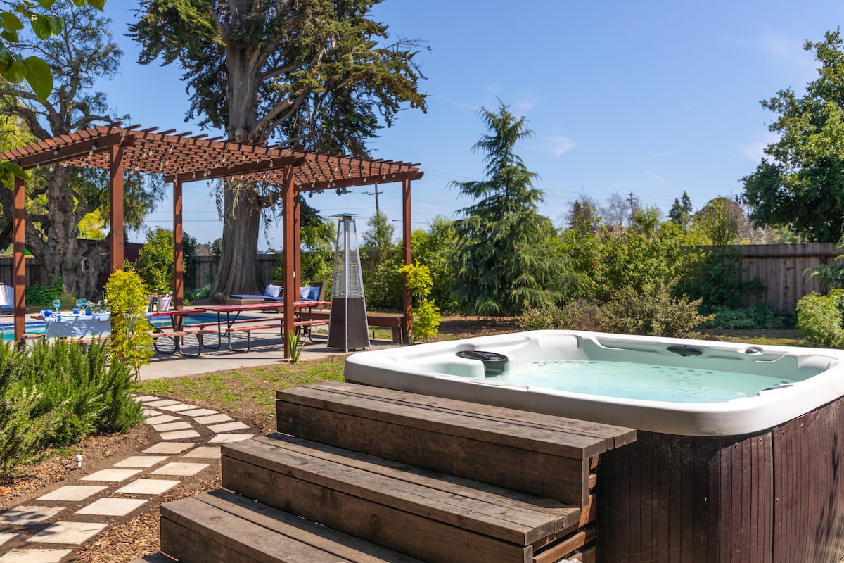 A hot tub is situated on a wooden deck, surrounded by lush greenery and vibrant plants. A pergola creates a shaded area nearby, while a spacious patio features dining areas in the background, all under a clear blue sky.