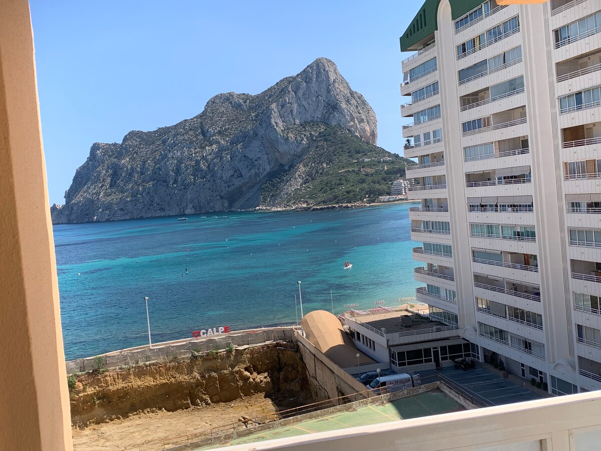 A view of the tranquil seaside features a rocky mountain backdrop, with vibrant blue waters gently lapping at the shore. A multi-story building stands nearby, complementing the scenic coastal landscape.