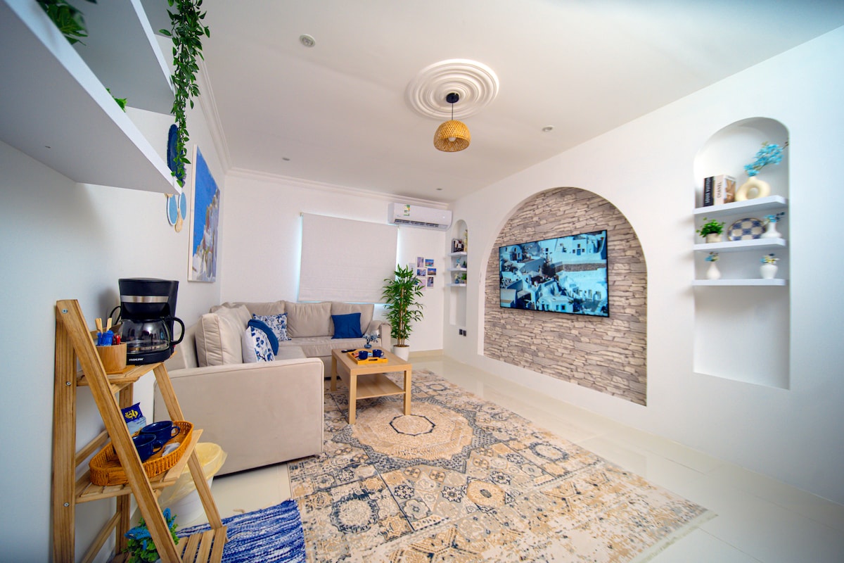 A comfortably arranged living area features a light-colored sofa and a wooden coffee table. A decorative stone wall is highlighted behind a flat-screen television. Natural light enters through a window, adding brightness to the space, complemented by plants and decorative shelves.