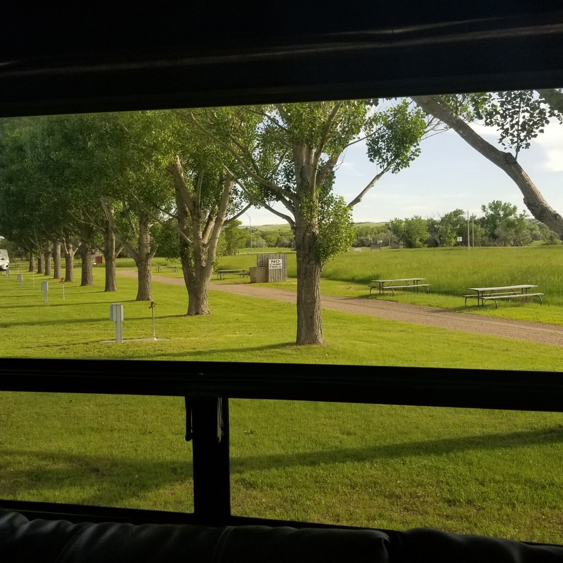 A view from inside an RV shows a tree-lined pathway leading to a facility. Lush green grass surrounds the area, with picnic tables visible in the distance. The scene offers a peaceful outdoor setting with ample natural light.