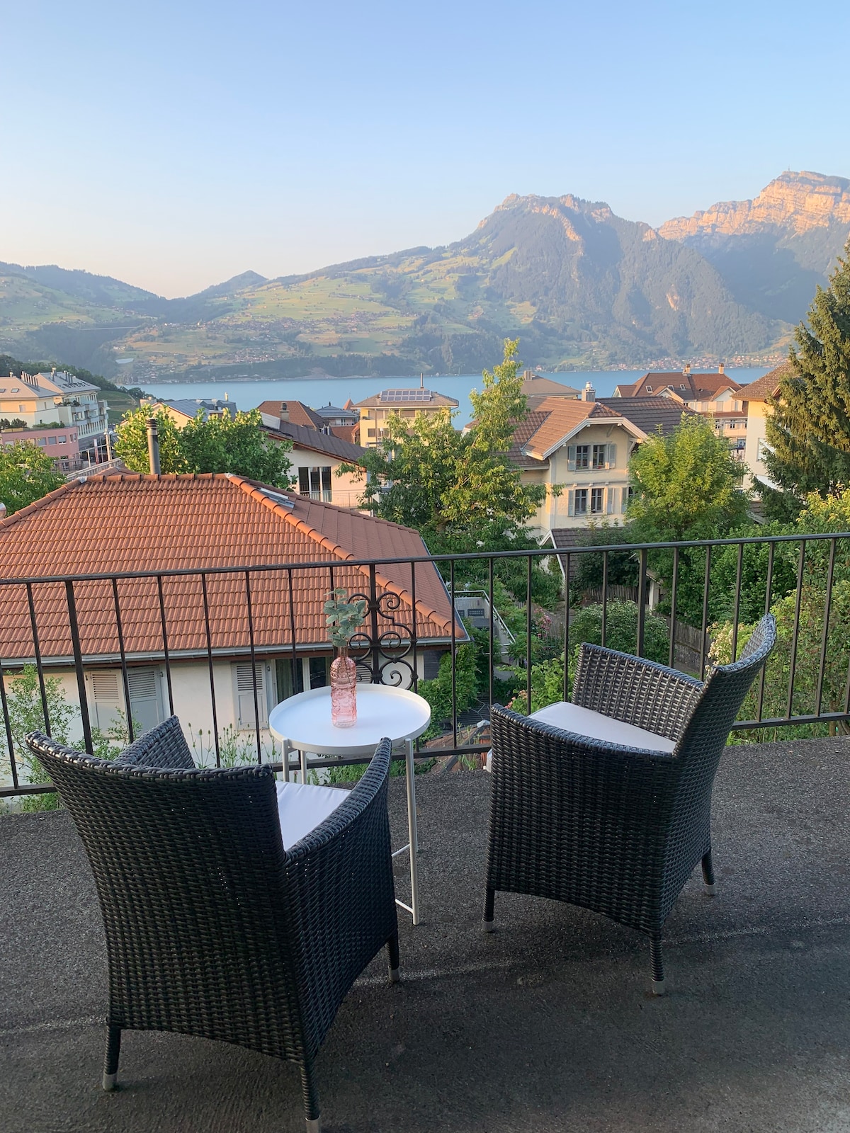A balcony seating area is presented with two wicker chairs positioned beside a small round table. The view includes lush mountains and a tranquil lake, framed by greenery and residential rooftops in the foreground.
