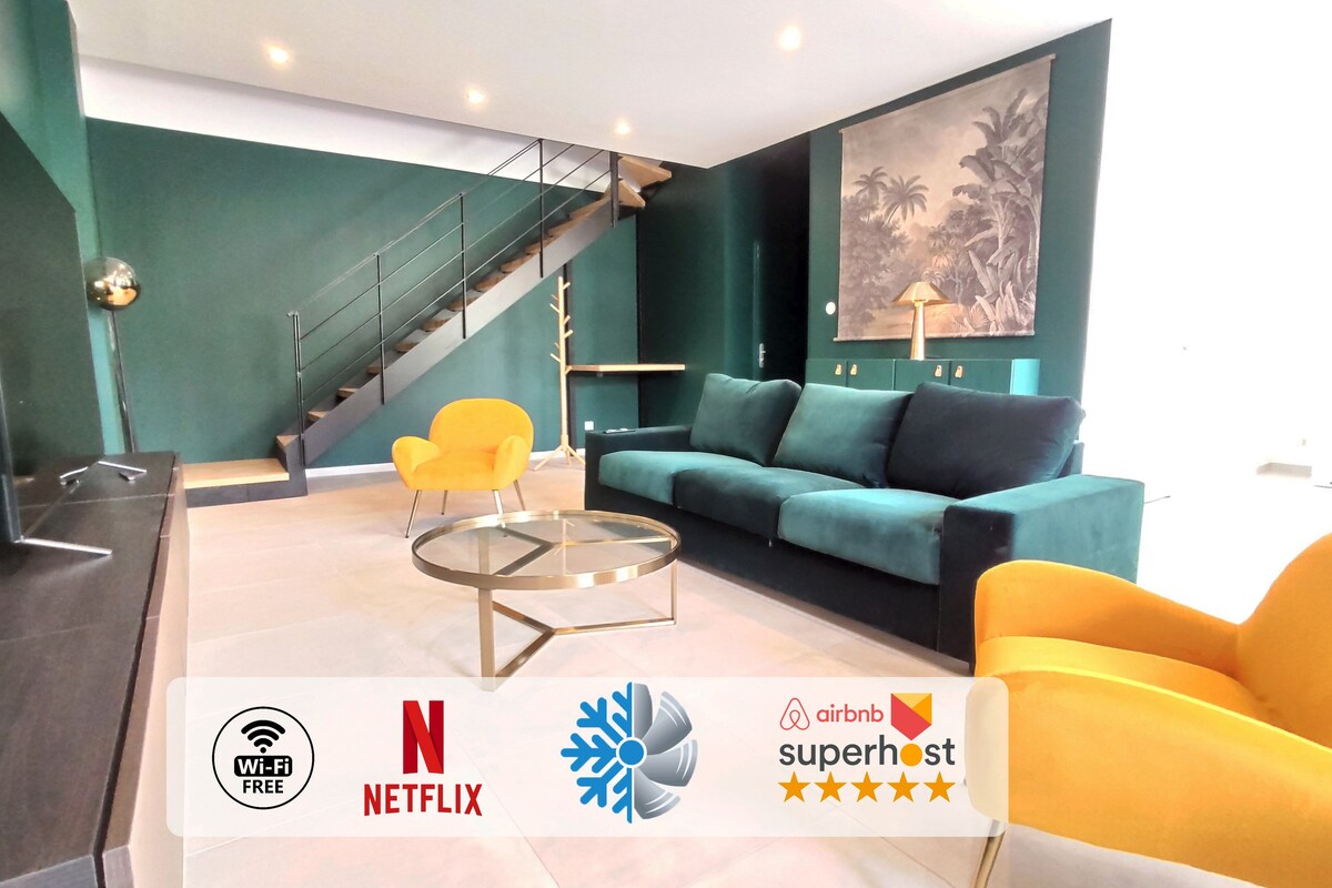 A spacious living area features deep green walls, a large sectional sofa in dark blue, and a glass coffee table. A staircase leads to an upper level, while a vibrant yellow chair adds a pop of color. A TV stand is positioned against the wall.