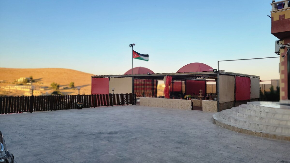 An outdoor setting features a spacious terrace with smooth stone flooring and a fenced perimeter. A gazebo with red canopies is visible, complemented by a backdrop of rolling hills. A flag representing Jordan can be seen fluttering in the breeze, enhancing the cultural atmosphere.