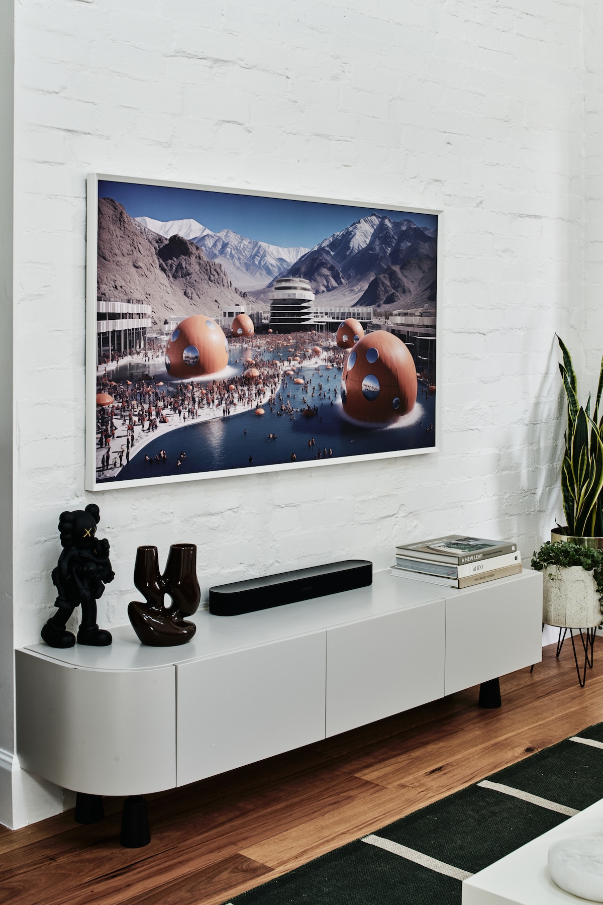 A sleek entertainment unit is positioned against a white wall, featuring a large framed artwork depicting a vibrant landscape with abstract structures. Two sculptures accompany an audio system, and a green plant adds a touch of nature to the modern design.