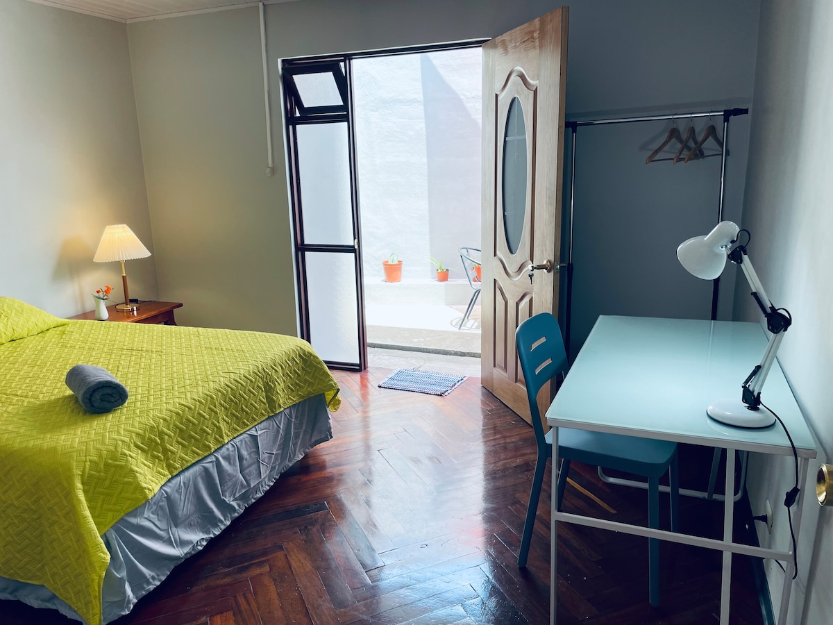 The sleeping area includes a spacious bed covered with a bright yellow quilt and a cozy bedside lamp. A desk with a lamp is positioned near the window, which opens to a private outdoor space. A clothing rack is also visible.