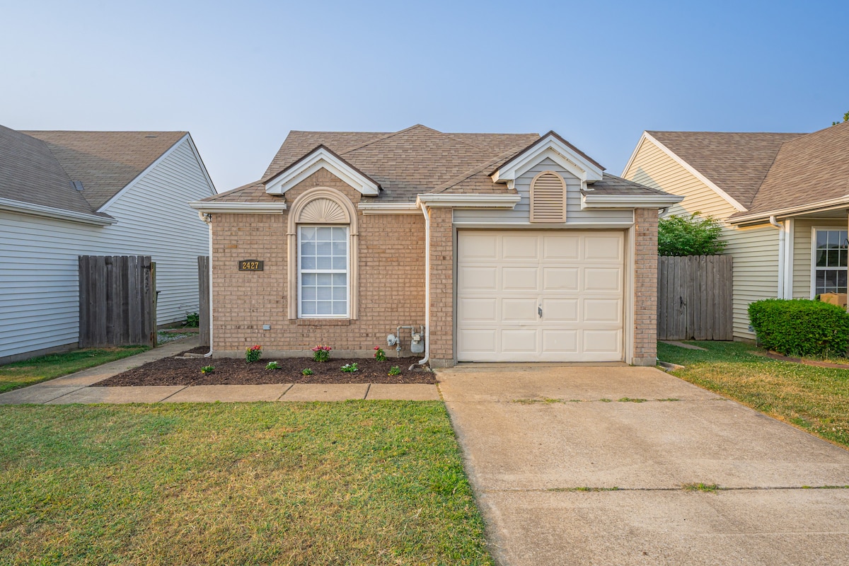 The exterior of a single-story residential home is shown, featuring a brick façade and a single-car garage. Neat landscaping includes small flowering plants in front, with a well-maintained lawn extending to the sidewalk. The neighboring homes are visible on either side.