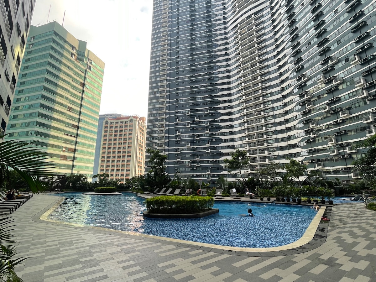 A well-maintained swimming pool is surrounded by modern condominium buildings, featuring landscaped areas with greenery. Lounge chairs are placed along the poolside, and the clear water reflects the surrounding architecture. The area is bathed in natural light, creating a serene atmosphere.