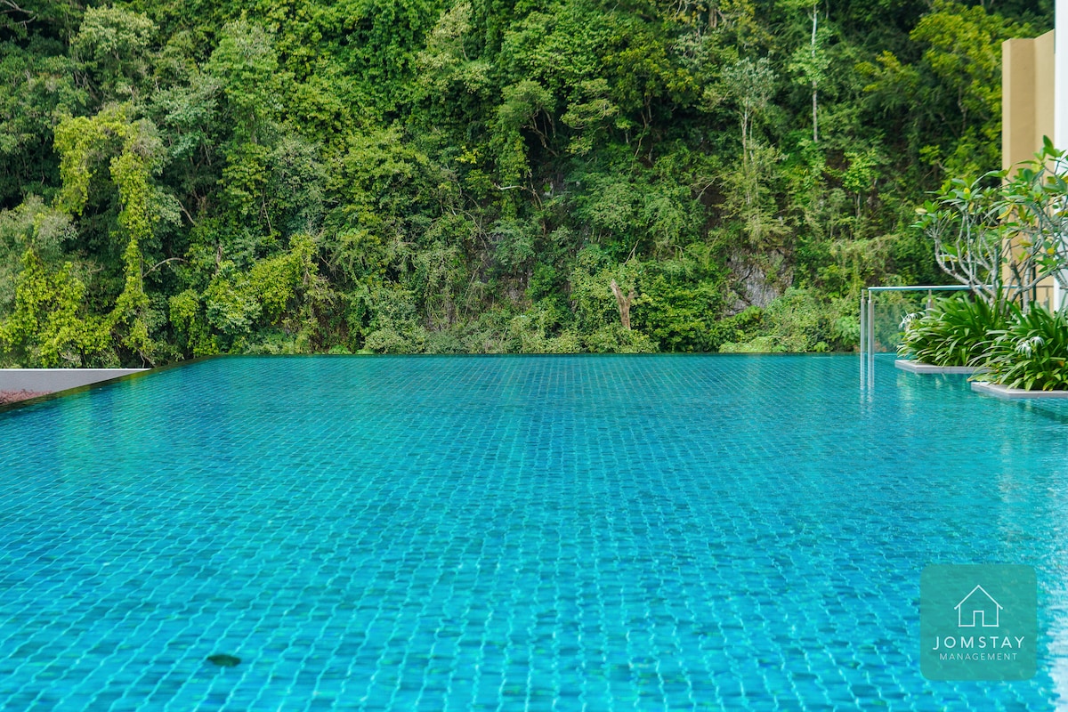 An infinity pool extends outward, featuring clear blue water that reflects the surrounding greenery. Lush foliage lines the backdrop, providing a natural setting. The pool's edge seamlessly blends into the view of the mountains, inviting relaxation and tranquility.