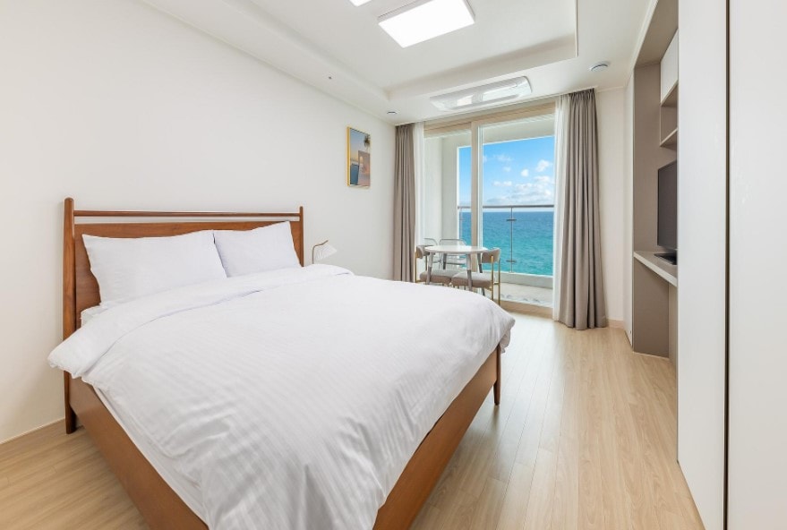 A spacious bedroom features a large bed with crisp white linens. A window offers a view of the ocean, while a small dining table and chairs are positioned nearby. Natural light fills the room through the window, creating a bright and airy feel.