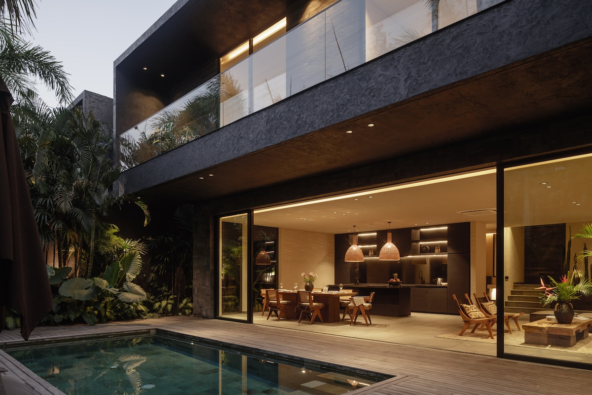A modern villa exterior showcases large glass doors that open to a spacious living area and kitchen. A wooden deck surrounds a swimming pool, while lush greenery enhances the serene atmosphere. The design combines indoor and outdoor spaces for an inviting and open feel.