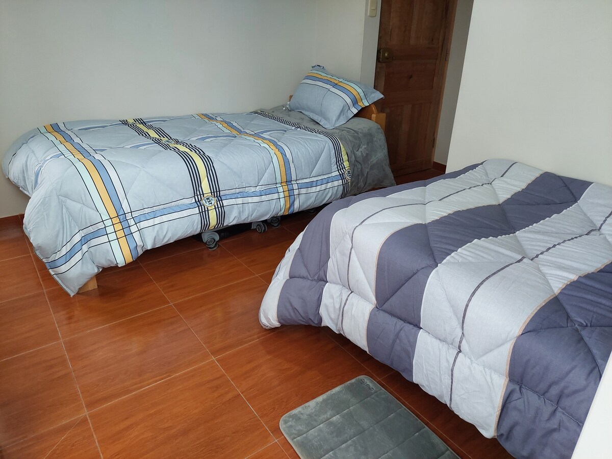 The sleeping area features two beds, each covered with patterned comforters. The flooring is a warm, polished wood, and a small mat is positioned beside the bed. The walls are painted a light color, enhancing the natural light in the room.