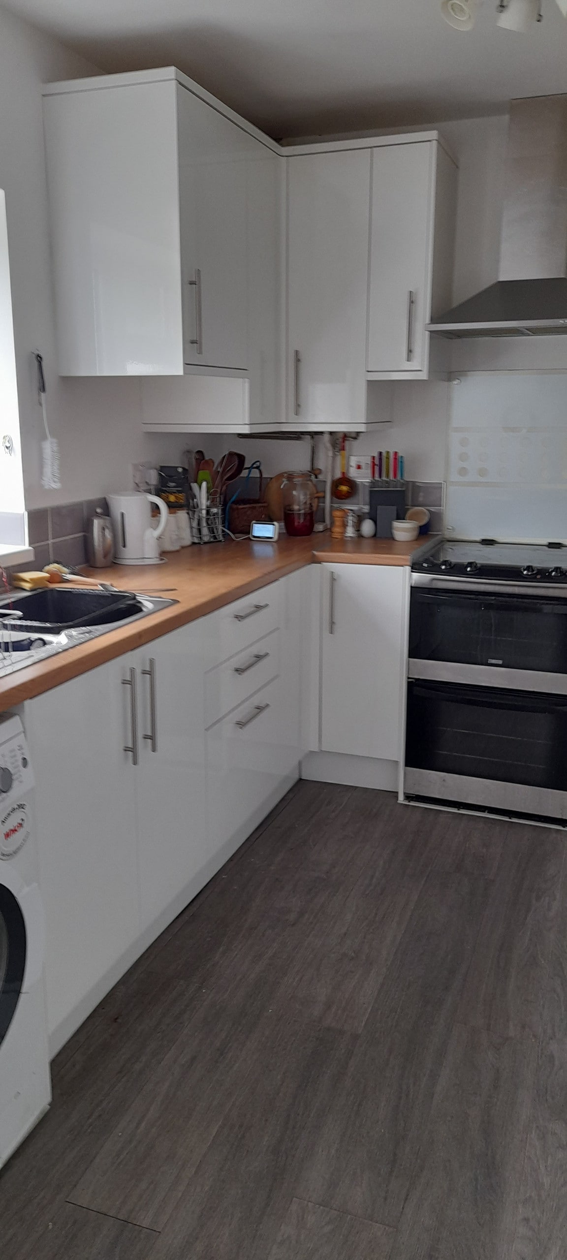 A modern kitchen is displayed with sleek white cabinetry and a wooden countertop. Cooking appliances, including a stove and oven, are visible along with utensils and dishware organized neatly. Natural light filters through the window, providing a bright and functional cooking space.